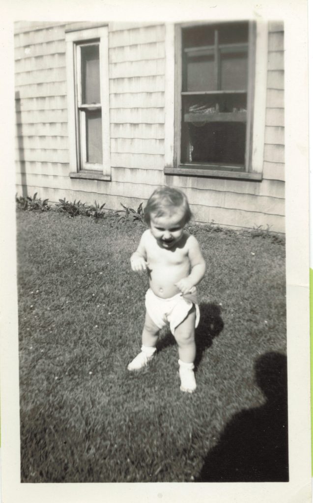 Janice Burrell Snyder about 1 year old. She's standing on grass next to a house.